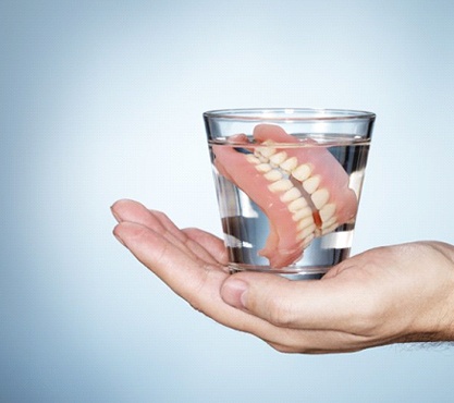 hand holding a glass of water with full dentures soaking in it